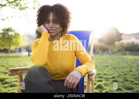Portrait de jeunes Afro-américaines attentionnés avec la main à la joue, regardant l'appareil photo tout en étant assis sur un transat dans un parc verdoyant, à la lumière du soleil Banque D'Images
