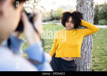 Récolte floue jeune homme photographe photographie de femmes afro-américaines debout près de l'arbre dans la pelouse avec la main derrière la tête tout en regardant CA Banque D'Images