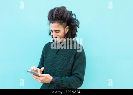 Jeune Afro-américain enthousiaste avec des cheveux bouclés dans un chandail décontracté parcourant un téléphone mobile tout en se tenant contre un fond de mur bleu Banque D'Images