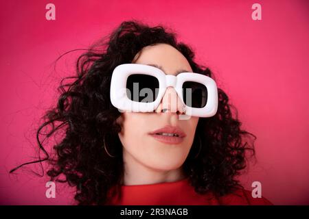Portrait de la jeune femme dans une tenue rouge tendance et de grandes lunettes de soleil blanches regardant l'appareil photo tout en se tenant sur fond rose dans le studio Banque D'Images