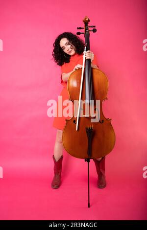 Corps complet de jeune musicienne ciblée avec des cheveux bouclés portant une robe brillante avec des bottes et regardant la caméra jouant du violoncelle en studio avec Banque D'Images
