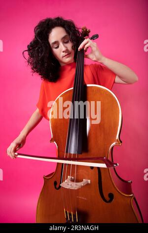 Corps complet de jeune musicienne ciblée avec des cheveux bouclés portant une robe brillante avec des bottes et regardant debout jouer au violoncelle en studio avec du rose Banque D'Images