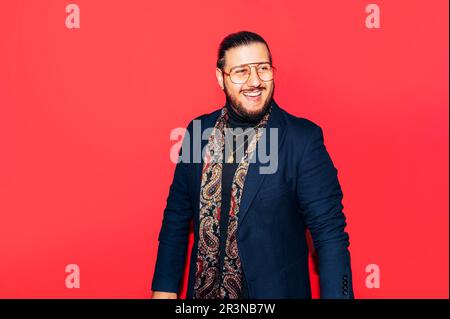Portrait d'un homme souriant et barbu en costume et lunettes tendance, sur fond rouge Banque D'Images