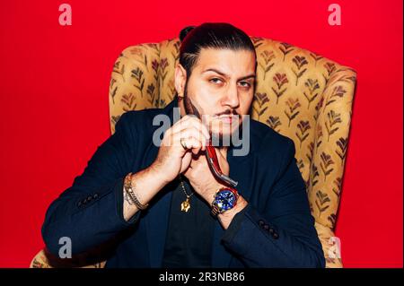 Homme brutal et sérieux avec une barbe portant des accessoires et une veste coûteux tout en regardant menaçant et en se rasant le visage avec un couteau tranchant sur fond rouge Banque D'Images