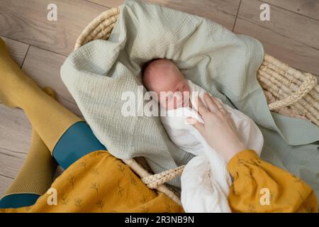 Vue de dessus de la femme méconnaissable se détendre sur un sol en bois à côté d'un lit de bébé en osier avec bébé dormant dans la selle blanche Banque D'Images