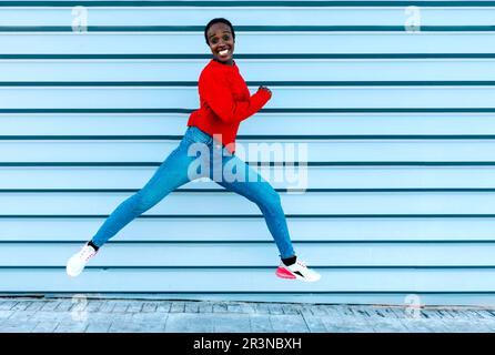 Vue latérale d'une femme noire excitée regardant l'appareil photo et souriant tout en faisant un saut contre le mur bleu sur la rue de la ville Banque D'Images