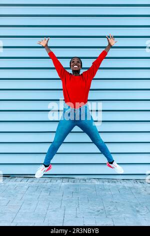 Tout le corps d'une femme afro-américaine joyeuse faisant un saut contre le mur bleu sur la rue de la ville tout en regardant vers le haut et souriant Banque D'Images