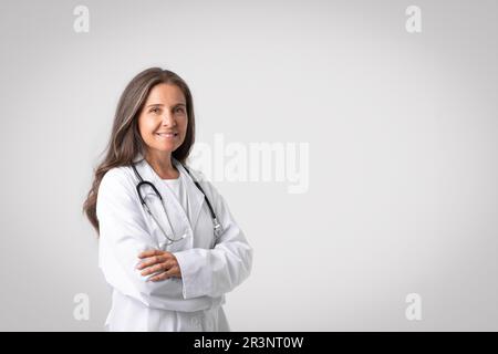 Portrait de gaie femme européenne senior médecin en manteau blanc avec stéthoscope debout avec bras pliés, espace copie Banque D'Images