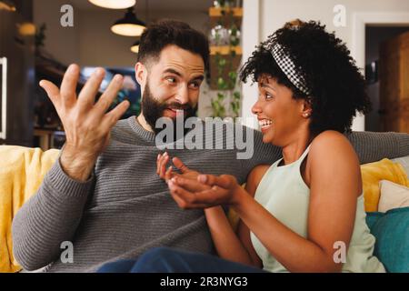 Joyeux jeune couple biracial regardant les uns les autres et parlant tout en étant assis sur un canapé dans la salle de séjour Banque D'Images