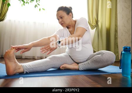 Femme gravid enceinte faisant des exercices d'étirement prénatal sur le tapis de fitness, pratiquant le yoga de grossesse à la maison. Banque D'Images