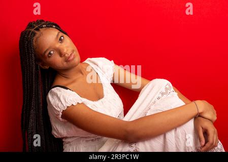 Belle jeune femme noire assise calmement vêtue d'une tenue blanche avec de longues tresses. Isolé sur fond rouge Banque D'Images