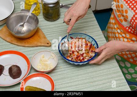 Gros plan sur la table de cuisine avec les mains de la femme qui organise tas de salade russe avec une cuillère dans un petit bol en plastique bleu vif. Banque D'Images