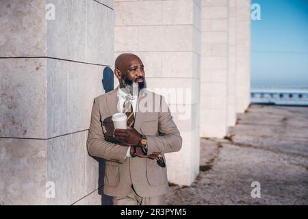 Un homme d'affaires africain attentionné se tient à l'extérieur près d'un pilier en calcaire, tenant une tasse de café et des lunettes de soleil. Il regarde loin de la caméra, avec le p Banque D'Images