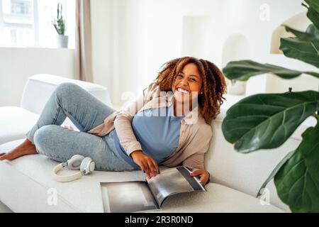 Une femme africaine assise sur un canapé à la maison tout en lisant un magazine ou un livre Banque D'Images