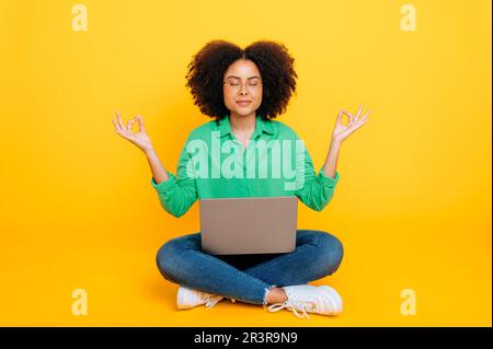Photo en longueur d'une femme curly afro-américaine ou brésilienne calme et détendue, élégamment habillée, assise sur un fond jaune avec un ordinateur portable, méditer avec les yeux fermés, prendre soin de la santé mentale, sourires Banque D'Images