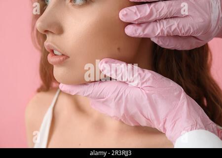 Dermatologue examinant les grains de beauté sur le visage de la jeune femme sur fond rose, gros plan Banque D'Images