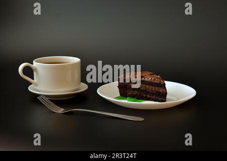 Un morceau de gâteau au chocolat avec des feuilles de menthe sur fond noir, et une tasse de café chaud sur une soucoupe avec une fourchette. Gros plan. Banque D'Images