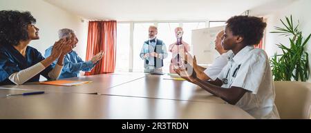 Dans un cliché panoramique, un médecin éminent avec des lunettes et une barbe nette, portant un gilet, se tient pour être applaudi par un groupe multiracial de col Banque D'Images