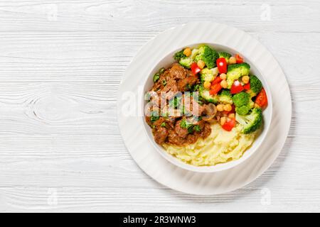 viande de bœuf hachée, ragoût de champignons servi avec une purée de pommes de terre et de pois chiches, tomates, brocolis, salade de fleurs dans un bol blanc sur une table en bois blanc, horizonta Banque D'Images