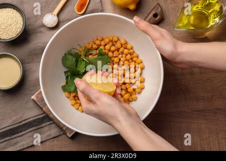 Femme pressant du jus de citron sur des pois chiches à une table en bois, vue de dessus. Cuisson de délicieux houmous Banque D'Images