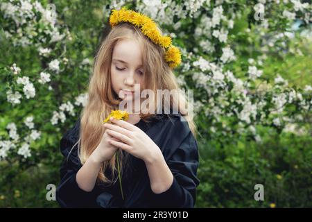 Une fille de 13 ans aux cheveux longs blonds et une couronne de pissenlits. Une fille blonde dans un jardin de fleurs de printemps cueillant des fleurs. La fille regarde Banque D'Images
