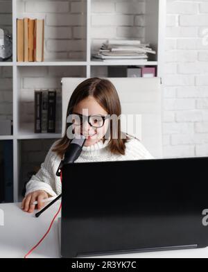 Petite fille dans des lunettes assis à la table blanche, enfant - blogueur souriant et parlant dans le microphone, enfant utilisant un ordinateur portable et regardant l'écran pendant r Banque D'Images