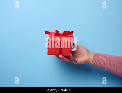 Une main de femme tenant une boîte cadeau rouge enveloppée de ruban sur fond bleu, un concept de félicitations, surprise Banque D'Images