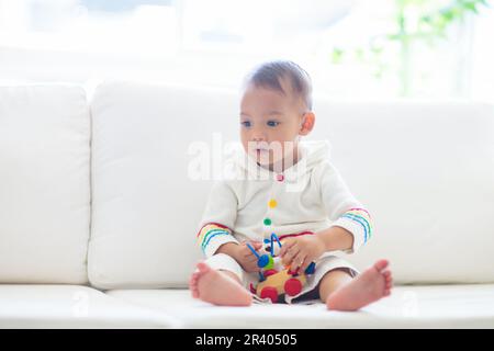 Bébé asiatique jouant avec un train de jouets coloré dans une chambre blanche ensoleillée. Bébé avec des jouets éducatifs. Enfant assis sur un canapé blanc. Banque D'Images