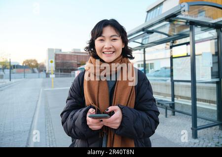 Portrait d'une femme coréenne en veste d'hiver, debout avec un smartphone, en attente d'un arrêt de bus, regardant l'application de téléphone mobile checki Banque D'Images