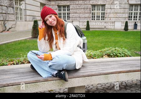 Portrait de jeune femme élégante, 25 ans, assis sur un banc dans le parc et utilise le téléphone mobile, lit les nouvelles en ligne, les messages ou les montres v Banque D'Images