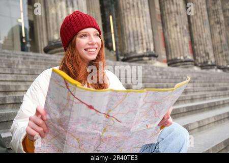 Portrait d'une jeune femme à tête rouge, le touriste se trouve avec une carte papier et cherche un itinéraire vers l'attraction touristique, repose sur les escaliers outdoo Banque D'Images