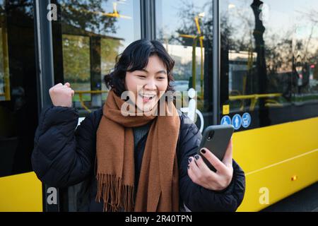 Portrait d'une jeune fille asiatique gaie parle sur téléphone mobile, chat vidéo, regarde stupéfié à l'appareil photo de smartphone, se trouve sur l'arrêt de bus Banque D'Images