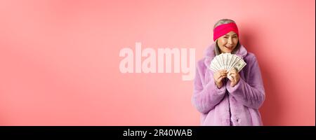 Shopping et mode. Femme asiatique à la mode pensant à l'achat de nouveaux vêtements, montrant l'argent en dollars et smi Banque D'Images