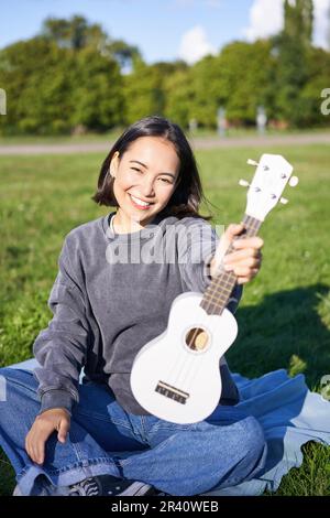 Musique et instruments. Une fille asiatique souriante montre son ukulele blanc, s'assoit dans le parc et joue une petite guitare Banque D'Images