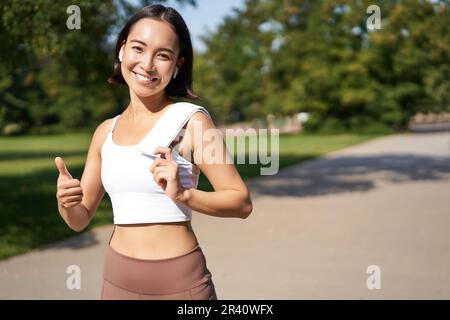 Une fille de forme physique souriante montre le pouce vers le haut, se tient avec une serviette après une bonne séance d'entraînement dans le parc, le jogging et l'air heureux Banque D'Images