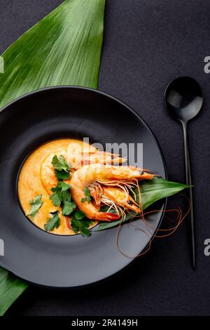 Soupe de fruits de mer à la crème avec crevettes décorées de feuilles tropicales. En plaque noire, sur fond noir. Vue de dessus, espace de copie. Banque D'Images