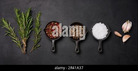 Des casseroles miniatures aux épices, au sel, au poivre noir et au poivre parfumé, une branche de romarin sur une table noire. Épices pour la cuisine Banque D'Images