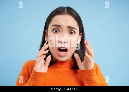 Gros plan de la femme asiatique choquée dans la panique, anxieux, semble terrifiée, se dresse sur fond bleu studio Banque D'Images