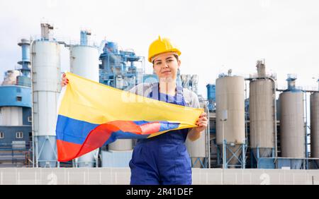 Une jeune femme ingénieure en casque agitant le drapeau de la Colombie tout en se tenant devant de grands réservoirs à l'usine chimique le jour ensoleillé du printemps Banque D'Images