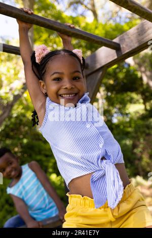 Vue à angle bas de l'afro-américain mignonne fille souriante accrochée sur les bars de singes à l'aire de jeux Banque D'Images