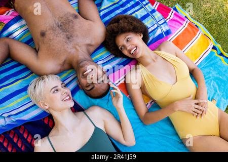 Joyeux groupe varié d'amis allongé sur des serviettes et souriant dans le jardin Banque D'Images