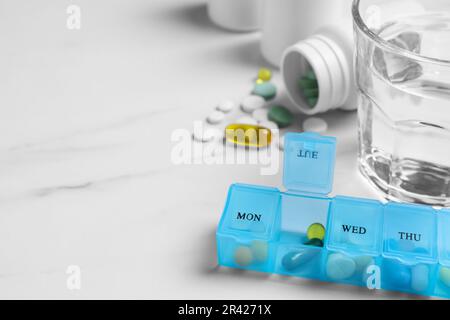 Boîte hebdomadaire avec médicaments et verre d'eau sur table en marbre blanc, gros plan. Espace pour le texte Banque D'Images