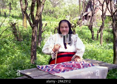 Comté de Maramures, Roumanie, 2000. Femme âgée en vêtements traditionnels à une table dans la cour. Banque D'Images