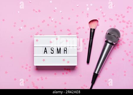 Microphone, pinceau et visionneuse de texte ASMR sur fond confetti rose. Concept de sons anti-stress d'enregistrement ASMR, pose plate, Banque D'Images