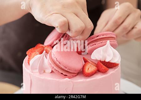 Femme pâtissière décorent le gâteau rose avec des macarons et des baies, gros plan.Processus de fabrication de gâteaux, mise au point sélective Banque D'Images