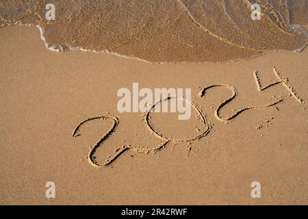 Concept de la nouvelle année. Texte 2024 manuscrit sur une surface de sable. La vague bleue de l'océan lavant les numéros à la plage. Année d'accueil 2024 bonne année prochaine de nouvelles résolutions, de nouveaux rêves et de nouveaux objectifs Banque D'Images