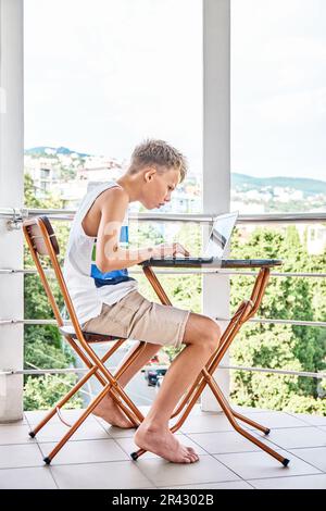 Préteen garçon avec l'expression sérieuse assis à la table regarde des vidéos sur ordinateur portable. L'élève aime regarder des vidéos pendant les vacances d'été à l'hôtel Banque D'Images