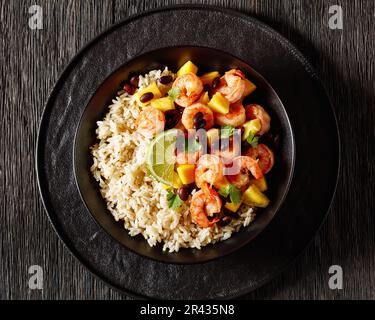 Crevettes grillées des Caraïbes aux haricots noirs, salsa à l'ananas à la mangue servie avec du riz brun dans un bol noir sur une table en bois sombre, vue horizontale depuis le dessus, Banque D'Images