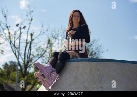 Roller blader femelle assis sur une rampe dans une piscine de patin en béton et de l'eau potable d'une bouteille de verre. Jeune patineuse fraîche portant un agresssiv moderne Banque D'Images