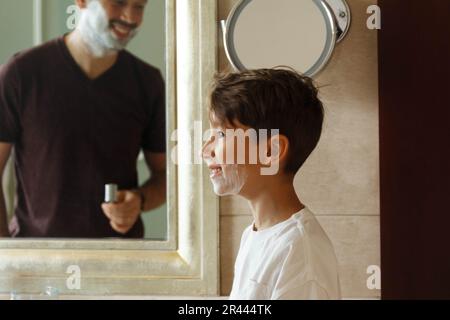 Un garçon avec de la mousse sur sa barbe regarde son père qui se rase Banque D'Images